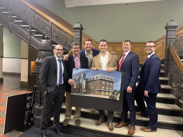Bruce Elia Jr. with his team on a grand staircase, holding a rendering of a new building development